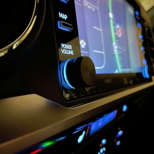 May include: A close-up of a car's dashboard with a black knob labelled "POWER VOLUME" and a blue backlight. The screen displays a map with a green line and a red dot.