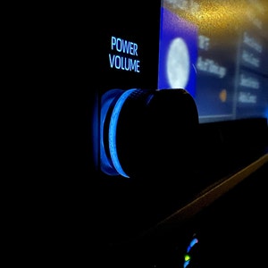 May include: A close-up of a car's radio volume knob with a blue backlight. The knob is labeled "POWER VOLUME".