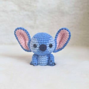 May include: A handmade crochet figure of a blue alien-like character with large ears and a dark blue nose. The inside of the ears is pink. The figure has black eyes and small arms and feet. The item is sitting on a white, fluffy surface.