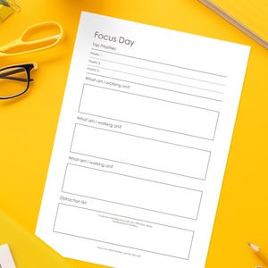 Pode incluir: Uma folha de papel branca intitulada "Focus Day" com seções para as principais prioridades e tarefas, em uma superfície amarela. Uma caneta, tesoura, óculos e uma planta também são visíveis.