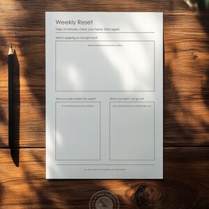 Op de afbeelding: Een witte Weekly Reset planner sheet met tekst en vakken voor journaling, op een houten oppervlak. De tekst bevat vragen als "Wat weegt er op me op dit moment?" en "Wat is er deze week echt belangrijk?" Een zwarte potlood ligt ernaast.