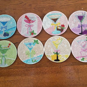 May include: Eight round drink coasters with cocktail designs. Each coaster features a different cocktail illustration, with colorful beaded details. The coasters are arranged on a wooden surface, showcasing a variety of drinks.