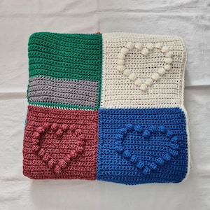 Könnte beinhalten: Ein handgehäkeltes Kissen mit Patchwork-Design. Das Kissen hat vier Quadrate in Grün, Weiß, Rot und Blau. Jedes farbige Quadrat hat ein Herzmuster aus Noppenstichen. Das Kissen ist ein gemütliches Wohnaccessoire.