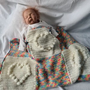 Könnte beinhalten: Handgefertigtes Häkel-Babydecken- und Wickelset. Die Decke hat ein großes weißes Herzdesign mit erhabenen Noppen und einen farbenfrohen Regenbogenrand. Die Wickel ist weiß mit Noppen und einem Regenbogenrand. Das Set ist für ein Baby konzipiert.