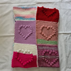 Könnte beinhalten: Eine gehäkelte Decke mit Patchwork-Muster, die aus verschiedenen farbigen Quadraten mit Herzmustern besteht. Die Decke ist in verschiedenen Farben gehalten, darunter Rosa, Lila, Rot, Grün und Weiß.