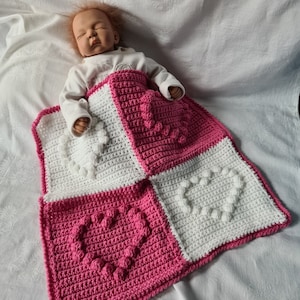Könnte beinhalten: Eine rosa-weiße gehäkelte Babydecke mit Herz-Design. Die Decke ist aus einem strukturierten Garn gefertigt und hat ein erhabenes Herzmuster.