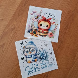 Könnte beinhalten: Zwei quadratische Mikrofasertücher mit gezackten Rändern. Ein Tuch zeigt eine Comic-Biene mit rotem Kopftuch, Blumen und dem Text "BEE happy". Das andere Tuch zeigt einen Pinguin mit blauer Mütze und Schal sowie den Text "Schön, dass es dich gibt".