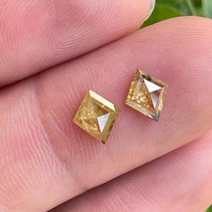May include: Two square-cut, yellow-toned diamonds. Each diamond has a faceted surface and a visible internal structure. The diamonds are held between fingers, with a blurred green background.