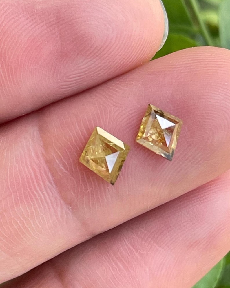 May include: Two square-cut, yellow-toned diamonds. Each diamond has a faceted surface and a visible internal structure. The diamonds are held between fingers, with a blurred green background.