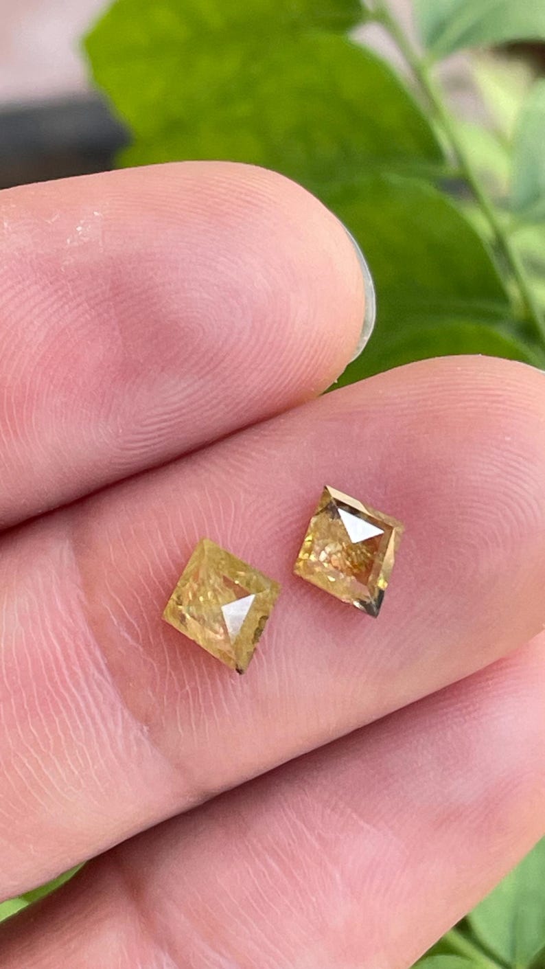 May include: Two square-cut, light yellow diamonds are displayed. The diamonds are held between fingers, showcasing their facets and clarity. The gems have a warm, golden hue, reflecting light.