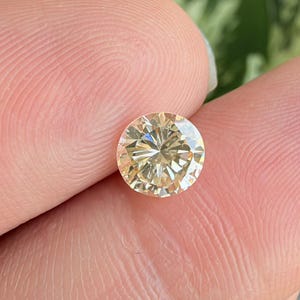 Pode incluir: Uma pedra preciosa redonda de corte brilhante com um tom champanhe claro ou amarelo pálido. A pedra apresenta numerosas facetas triangulares e em forma de pipa que criam um padrão complexo de reflexão e refração da luz na superfície.