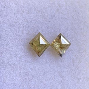May include: Two square-cut, light yellow diamonds. Each diamond has a flat top and a pointed bottom, with facets that catch the light. The diamonds are set against a soft, white background.