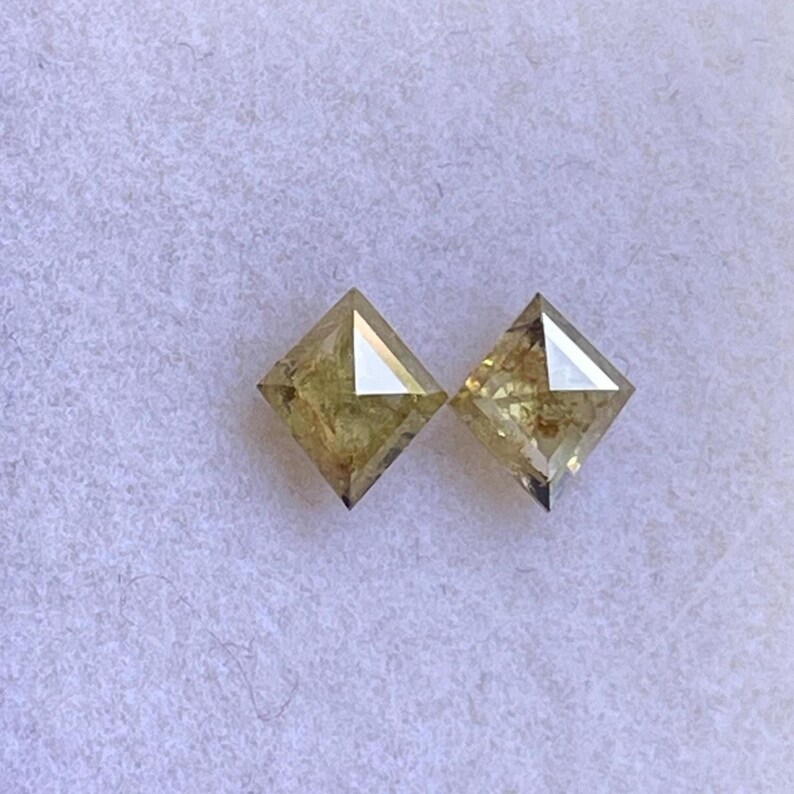 May include: Two square-cut, light yellow diamonds. Each diamond has a flat top and a pointed bottom, with facets that catch the light. The diamonds are set against a soft, white background.