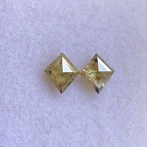 May include: Two square-cut, light yellow diamonds. Each diamond has a flat top and a pointed bottom. The diamonds are set against a white, textured background. The diamonds are likely for jewelry.