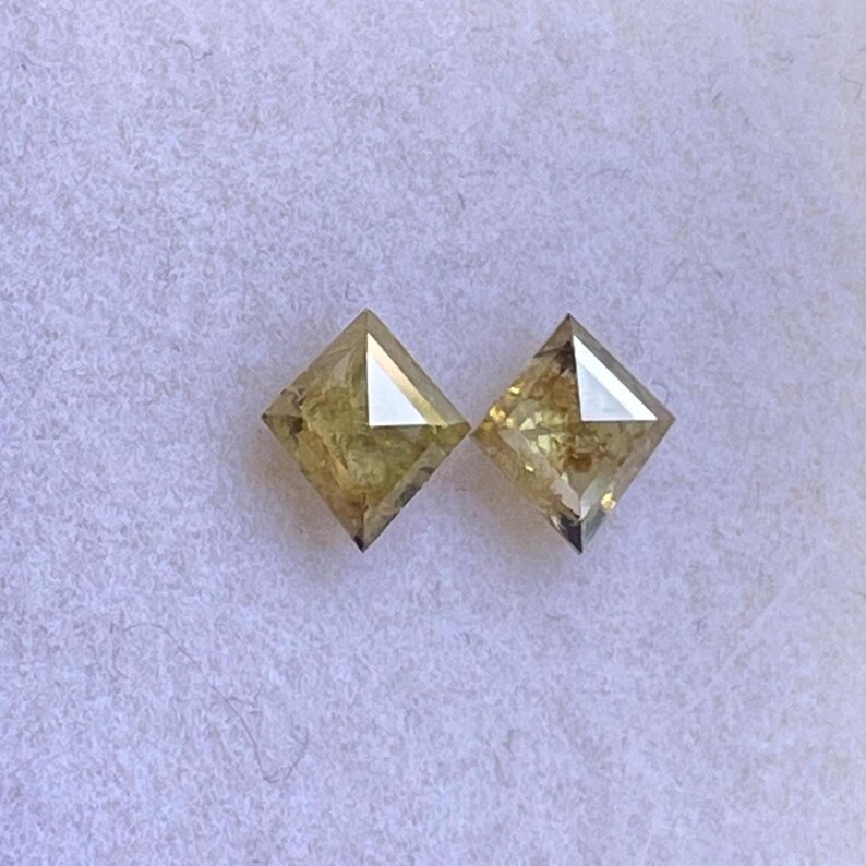 May include: Two square-cut, light yellow diamonds. Each diamond has a flat top and a pointed bottom. The diamonds are set against a white, textured background. The diamonds are likely for jewelry.