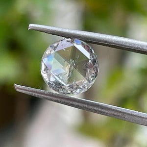 May include: A round, clear diamond held by metal tweezers. The diamond has multiple facets that reflect light, creating a rainbow effect. The background is a blurred green.