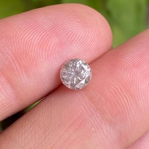 May include: A round, faceted diamond with a light gray hue. The gemstone is held between two fingers, showcasing its brilliance and clarity. The diamond's facets reflect light, creating a sparkling effect. This image highlights the beauty of a single gemstone.