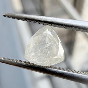 May include: A loose, trillion-cut diamond with a translucent, off-white appearance and visible internal inclusions. The gemstone features a triangular shape with faceted surfaces that reflect light, held securely by metal tweezers.