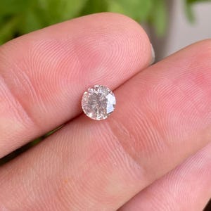 May include: A round, faceted diamond with a light champagne color. The gemstone is held between fingers, showcasing its brilliance and clarity. The diamond's facets reflect light, creating a sparkling effect, perfect for jewelry.