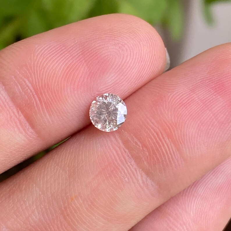 May include: A round, faceted diamond with a light champagne color. The gemstone is held between fingers, showcasing its brilliance and clarity. The diamond's facets reflect light, creating a sparkling effect, perfect for jewelry.