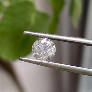 May include: A round, clear diamond held by silver tweezers. The diamond has many facets that reflect light, creating a sparkling effect. The background is blurred, with green foliage visible.