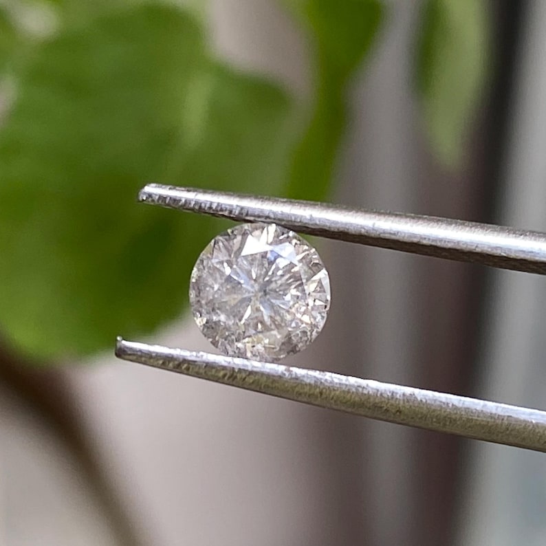 May include: A round, clear diamond held by silver tweezers. The diamond has many facets that reflect light, creating a sparkling effect. The background is blurred, with green foliage visible.