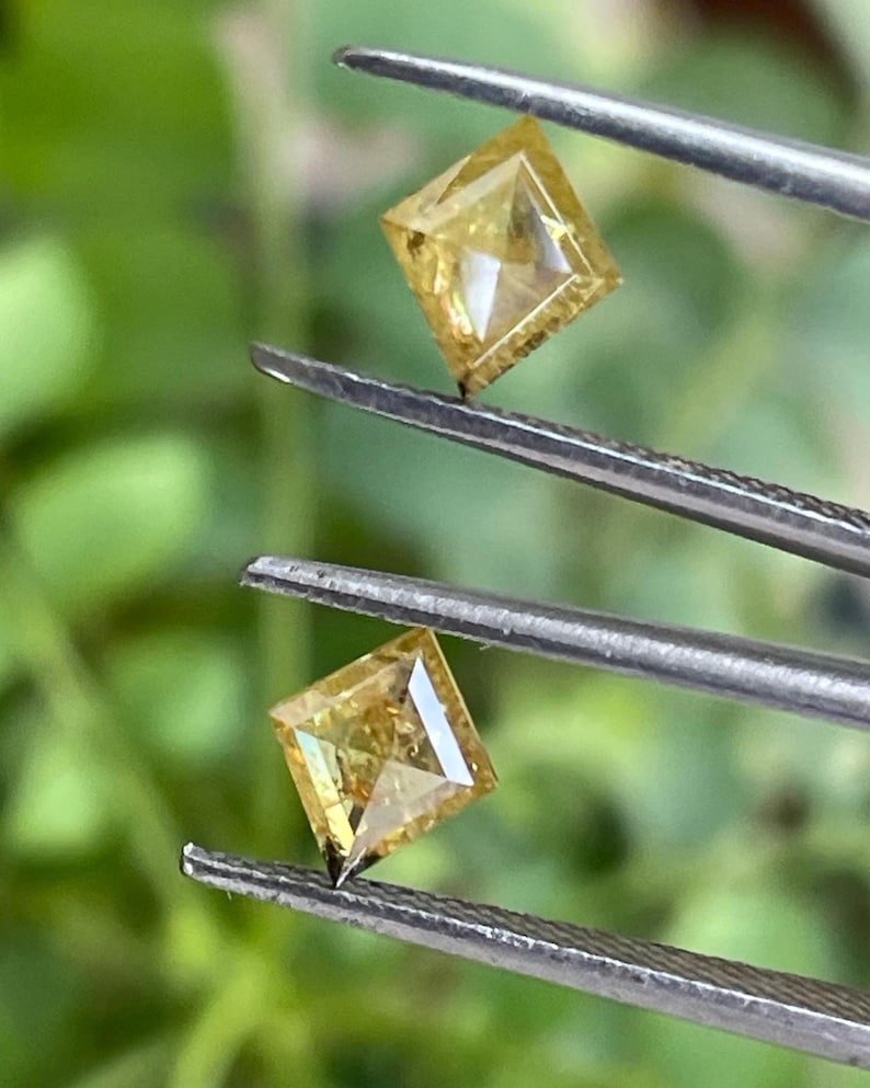 May include: Two square-cut, yellow diamonds are held by tweezers against a blurred green background. The diamonds have a brilliant cut, reflecting light. The tweezers are silver-colored.