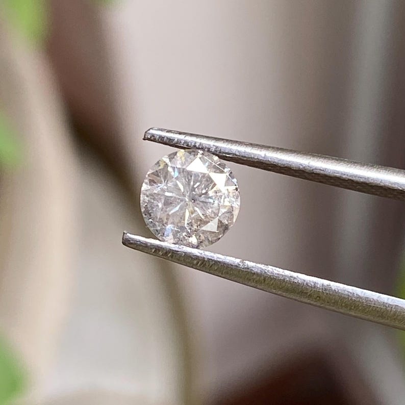 May include: A round, clear diamond held by silver tweezers. The diamond has multiple facets that reflect light, creating a sparkling effect. The background is blurred, with hints of green and beige.