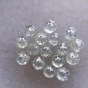 May include: A collection of clear, faceted gemstones. The round-cut stones are scattered across a light-colored surface, each reflecting light with multiple facets. The gems vary slightly in size and clarity, creating a sparkling display.