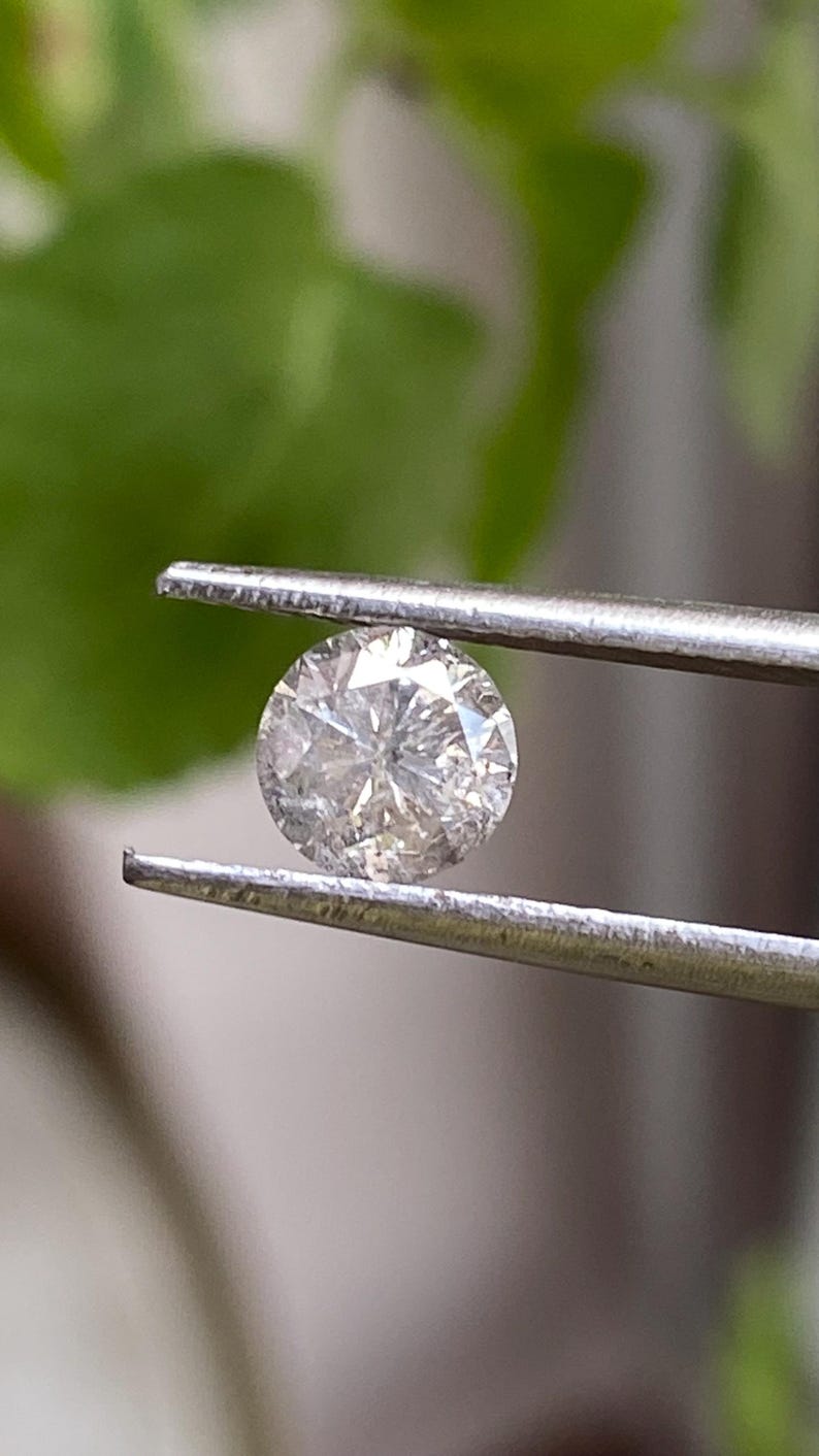 May include: A round, brilliant-cut diamond held by a pair of silver tweezers. The diamond sparkles with internal facets, reflecting light. The background is blurred green foliage.