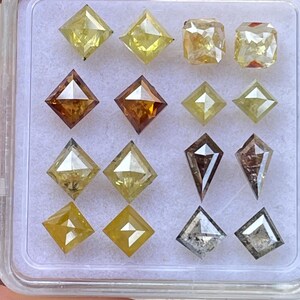 May include: A collection of various-shaped diamonds in a clear plastic container. The diamonds are in shades of yellow, brown, and gray. Shapes include square, kite, and rectangular cuts. The diamonds are displayed on a white surface.