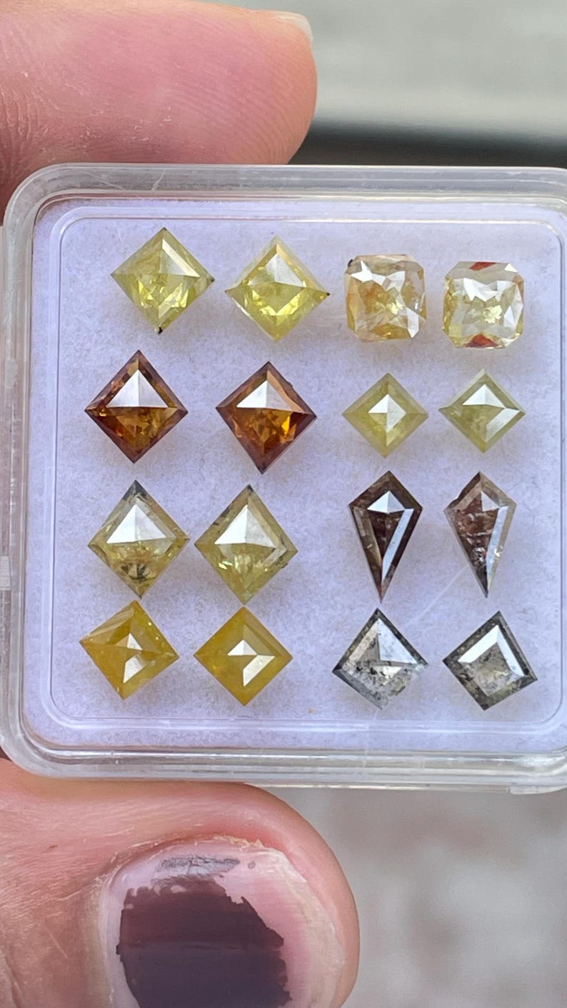 May include: A collection of various-shaped diamonds in a clear plastic container. The diamonds are in shades of yellow, brown, and gray. Shapes include square, kite, and rectangular cuts. The diamonds are displayed on a white surface.
