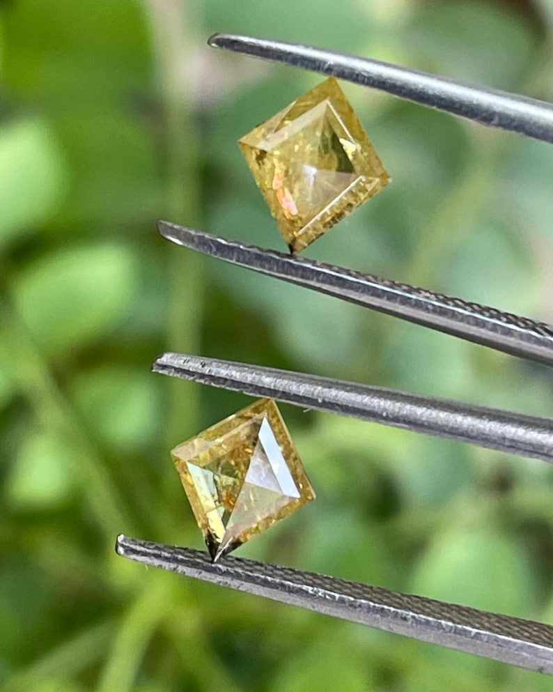 May include: Two square-cut, yellow diamonds are held by tweezers against a blurred green background. The diamonds have a brilliant cut, reflecting light. The gems are likely for jewelry making.