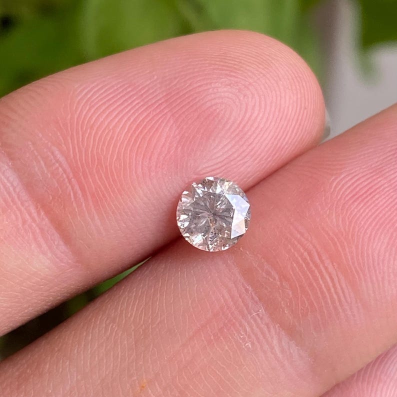 May include: A round, light brown diamond with multiple facets. The gemstone is held between fingers, with a blurred green background. The diamond's cut allows light to reflect, creating a sparkling effect.