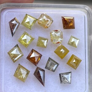 May include: A collection of loose, cut diamonds in various shapes and colors, including square, kite, and rectangular forms. The diamonds range in color from clear to yellow, brown, and gray, displayed in a clear plastic container.