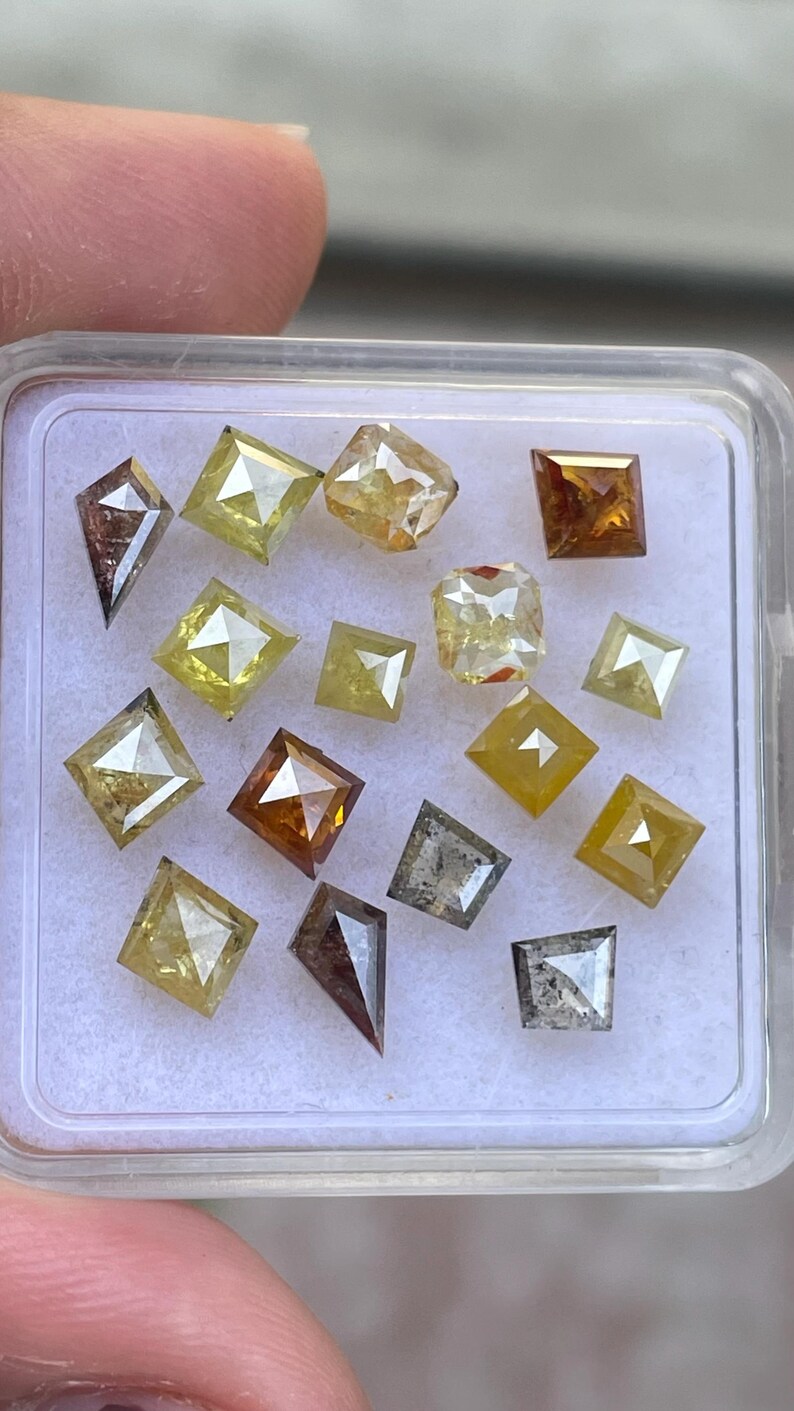May include: A collection of loose, cut diamonds in various shapes and colors, including square, kite, and rectangular forms. The diamonds range in color from clear to yellow, brown, and gray, displayed in a clear plastic container.