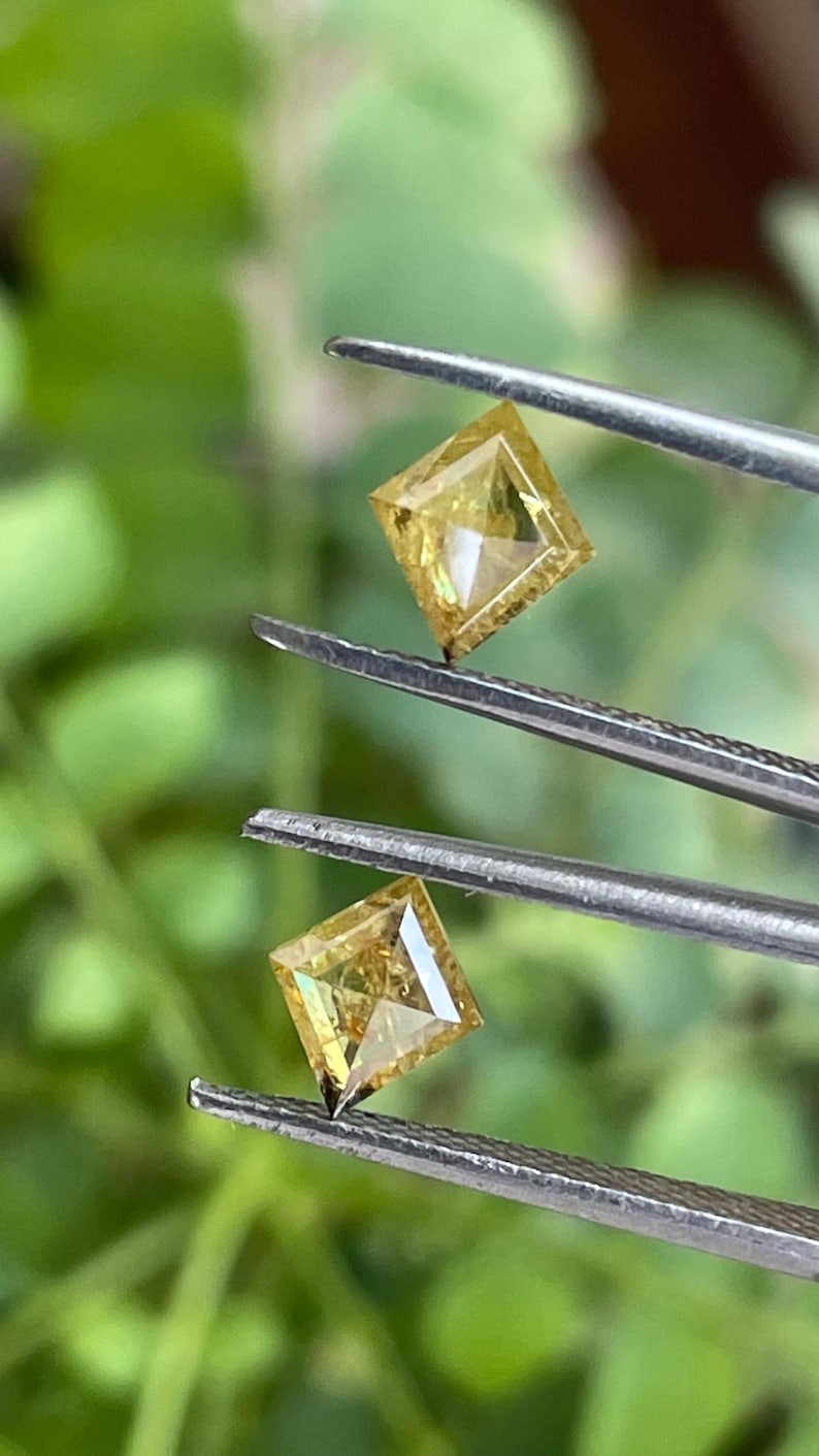 May include: Two square-cut, yellow diamonds are held by tweezers against a blurred green background. The diamonds have a bright, golden hue and are precisely cut with visible facets, showcasing their brilliance. The tweezers are silver.