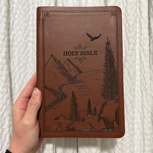 Puede incluir: Una Biblia de tapa dura de cuero marrón con un diseño en relieve de montañas, un río y un pájaro volando sobre las montañas. El texto "HOLY BIBLE" está en relieve en la portada.