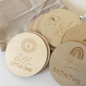 Pu&ograve; includere: Un set di dischi in legno per le tappe fondamentali del bambino con disegni e testo incisi. I dischi presentano illustrazioni di un leone, un elefante e un arcobaleno, insieme alle parole "hello LITTLE ONE", "three" e "two MONTHS". &Egrave; visibile un sacchetto beige con coulisse.