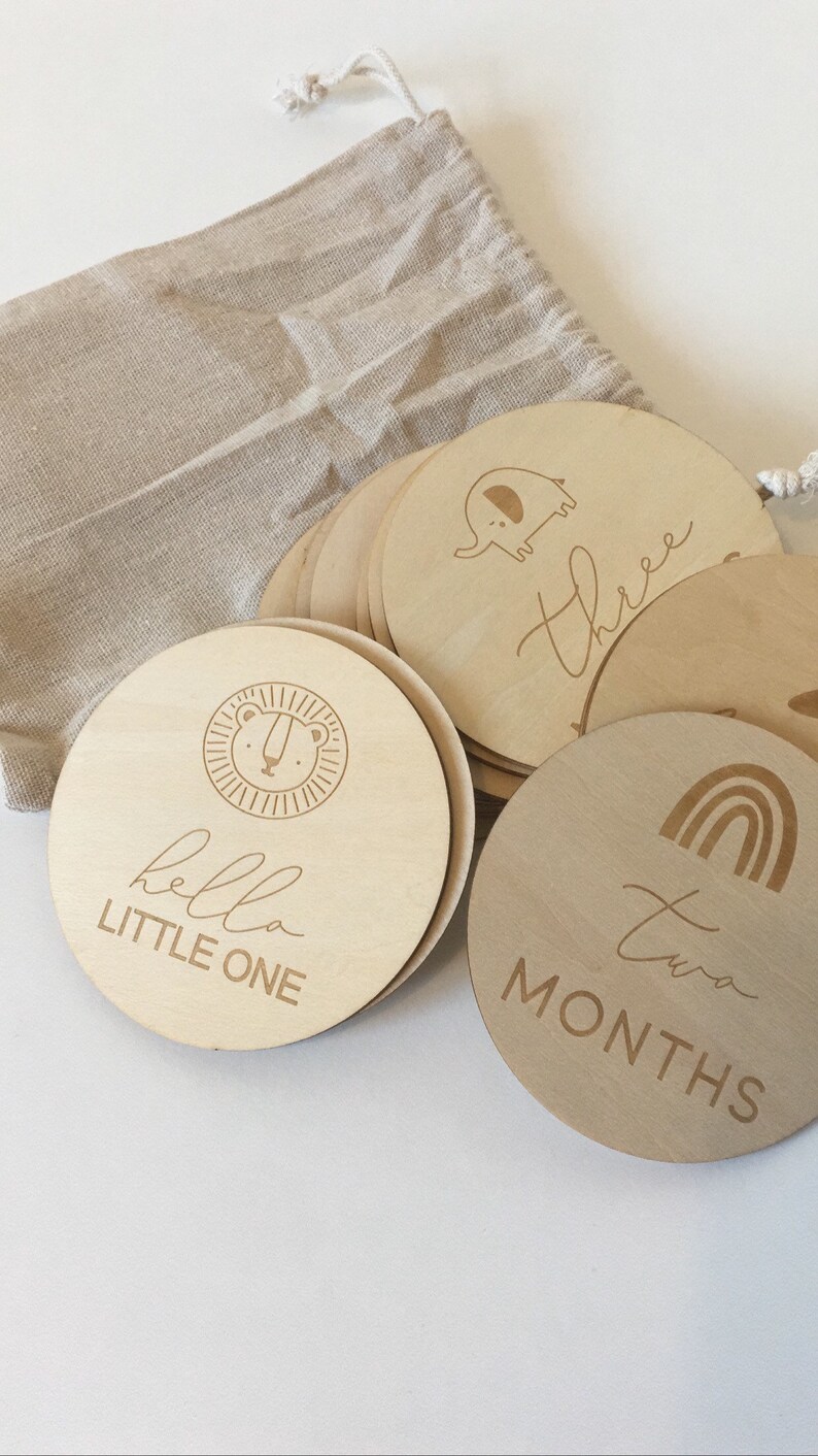 Pu&ograve; includere: Un set di dischi in legno per le tappe fondamentali del bambino con disegni e testo incisi. I dischi presentano illustrazioni di un leone, un elefante e un arcobaleno, insieme alle parole "hello LITTLE ONE", "three" e "two MONTHS". &Egrave; visibile un sacchetto beige con coulisse.