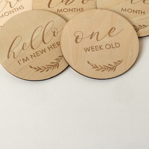 Pu&ograve; includere: Una collezione di dischi di legno rotondi per pietre miliari con testo inciso. I dischi presentano numeri e frasi come "una settimana" e "sono nuovo qui", insieme a dettagli di foglie decorative. I dischi sono marroni chiari.