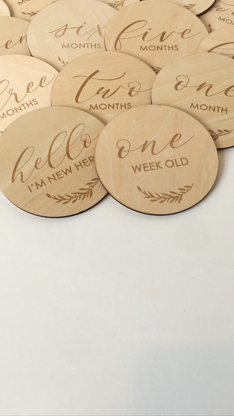 Pu&ograve; includere: Una collezione di dischi di legno rotondi per pietre miliari con testo inciso. I dischi presentano numeri e frasi come "una settimana" e "sono nuovo qui", insieme a dettagli di foglie decorative. I dischi sono marroni chiari.