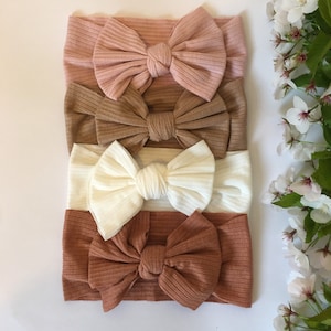 May include: Four different coloured ribbed headbands with bows. The headbands are in shades of pink, brown, white, and orange.