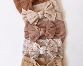 Può includere: Cinque diversi scrunchies per capelli con un design a fiocco annodato. Gli scrunchies sono in tonalità di marrone, beige e bianco. Uno scrunchie ha un motivo floreale.