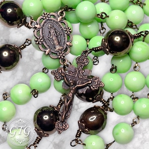 May include: A rosary featuring light green beads and bronze-colored metal accents. The rosary includes a crucifix and a medallion with religious imagery. The beads are round and evenly spaced, with larger, darker beads interspersed throughout.