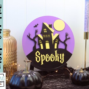 May include: Purple and black Halloween-themed round sign with a silhouette of a spooky house and the word "Spooky" in yellow letters.