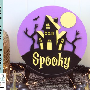 May include: Purple and black Halloween sign with a spooky house and a full moon. The sign says "Spooky" in a black font.