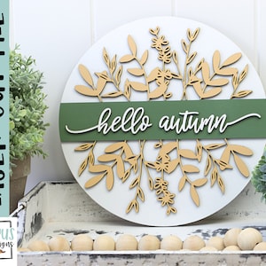 May include: A round, white wooden sign with laser-cut botanical elements in natural wood tones. A green band across the center displays the words "hello autumn" in white script. The sign is a laser cut file.