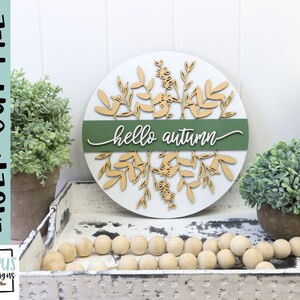 May include: A round, white wooden sign with the words "hello autumn" in cursive. The sign features laser-cut leaf and floral designs in a light brown color, with a green band across the center. The words "laser cut file" are printed vertically on the left.