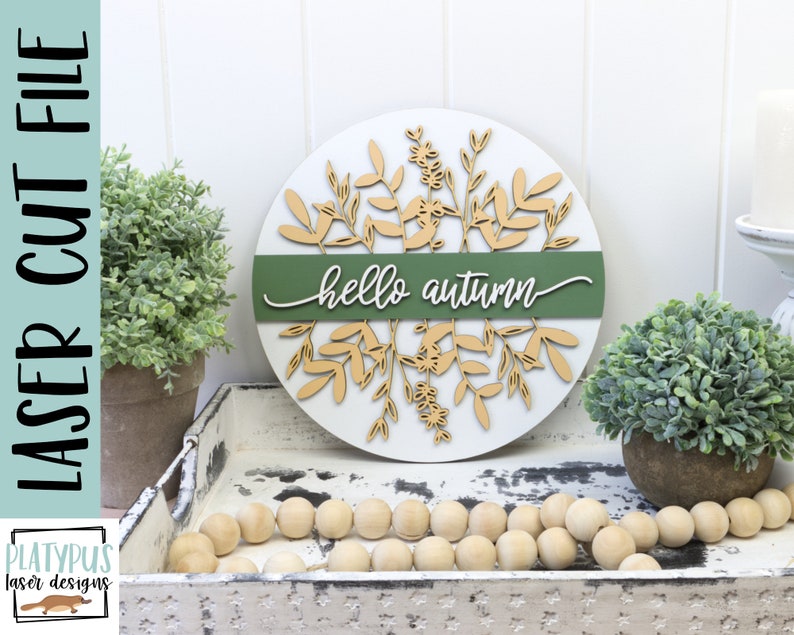 May include: A round, white wooden sign with the words "hello autumn" in cursive. The sign features laser-cut leaf and floral designs in a light brown color, with a green band across the center. The words "laser cut file" are printed vertically on the left.